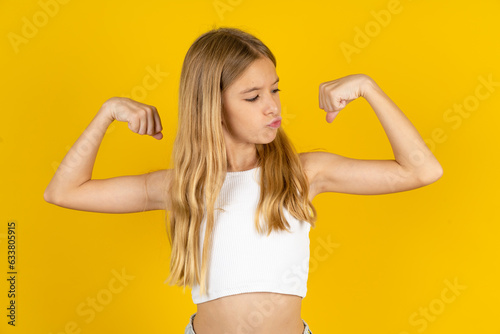 blonde kid girl wearing white T-shirt over yellow studio background showing arms muscles smiling proud. Fitness concept.