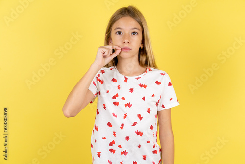 blonde kid girl wearing polka dot shirt over yellow studio background mouth and lips shut as zip with fingers. Secret and silent, taboo talking.
