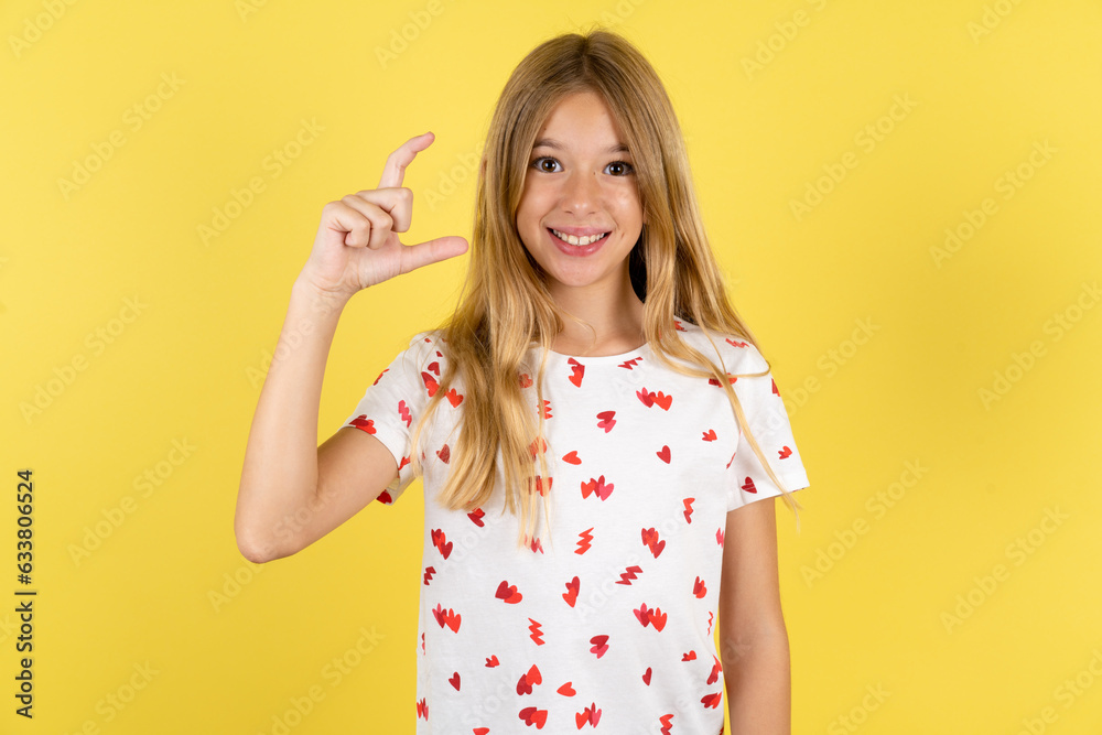 blonde kid girl wearing polka dot shirt over yellow studio background smiling and confident gesturing with hand doing small size sign with fingers looking and the camera. Measure concept blonde kid girl wearing polka dot shirt over yellow studio background smiling and confident gesturing with hand doing small size sign with fingers looking and the camera. Measure concept