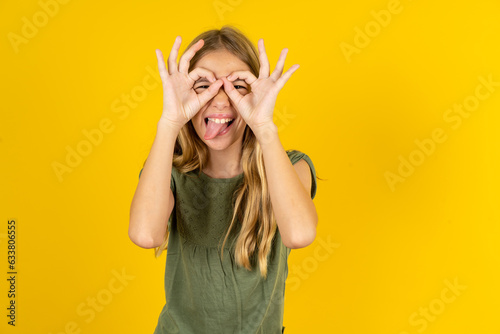 blonde kid girl wearing green T-shirt over yellow studio background doing ok gesture like binoculars sticking tongue out, eyes looking through fingers. Crazy expression.