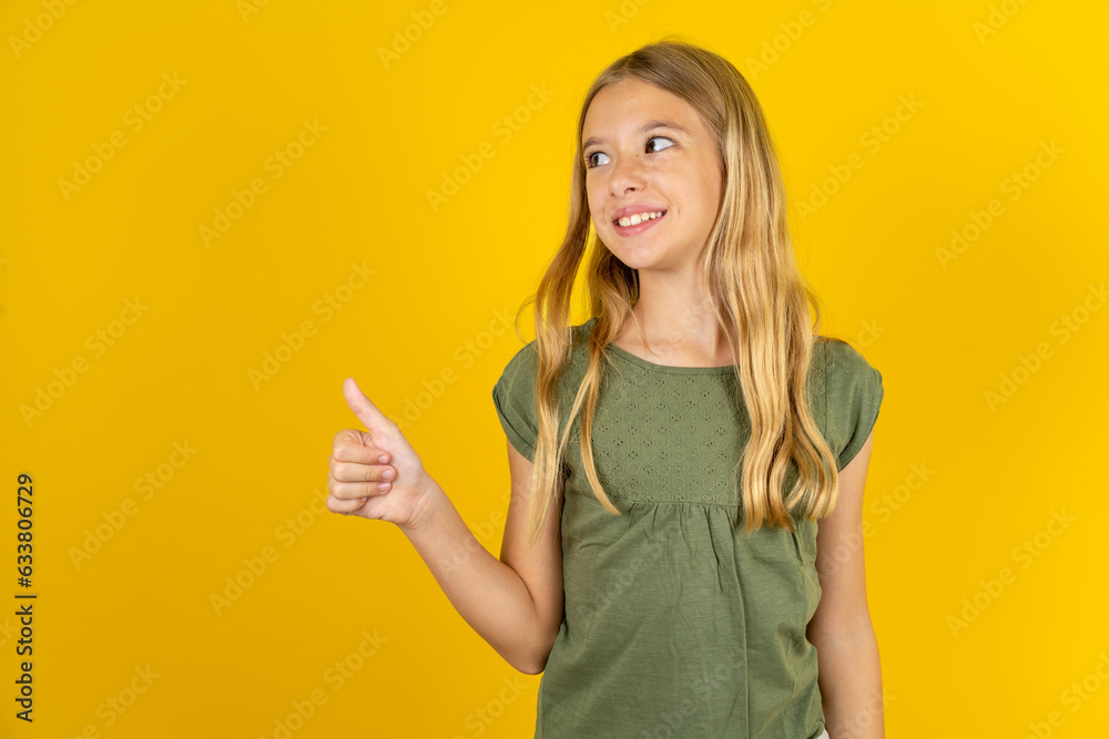 blonde kid girl wearing green T-shirt over yellow studio background Looking proud, smiling doing thumbs up gesture to the side. Good job!
