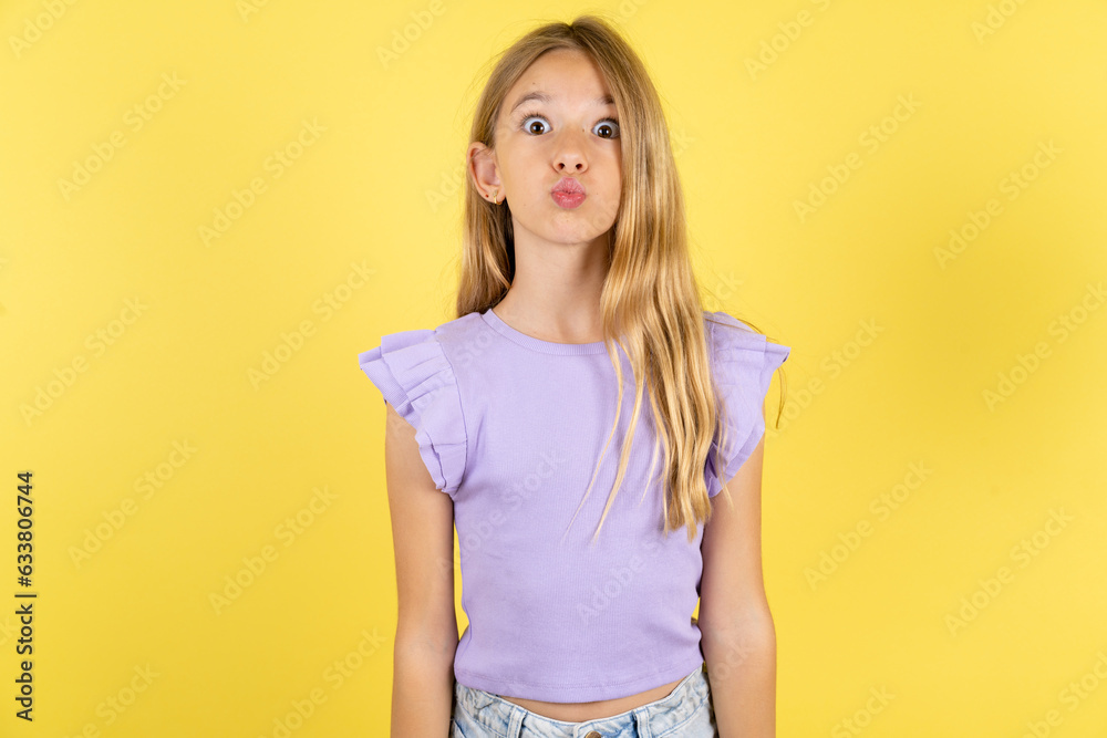 Shot of pleasant looking blonde kid girl wearing violet T-shirt over yellow studio background , pouts lips, looks at camera, Human facial expressions Shot of pleasant looking blonde kid girl wearing violet T-shirt over yellow studio background , pouts lips, looks at camera, Human facial expressions