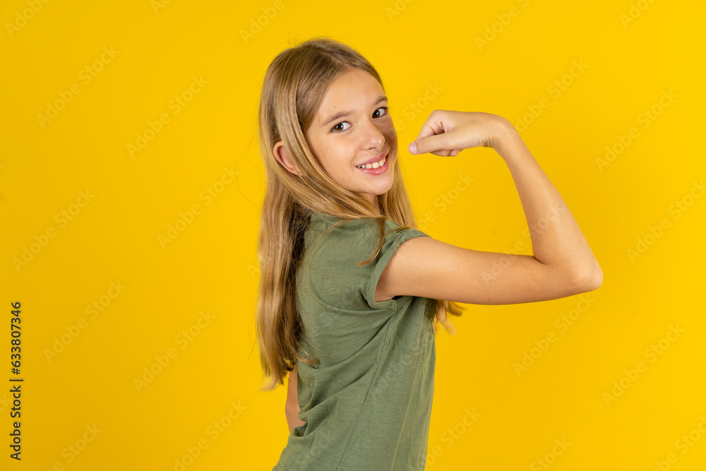 Portrait of powerful cheerful blonde kid girl wearing green T-shirt over yellow studio background showing muscles.