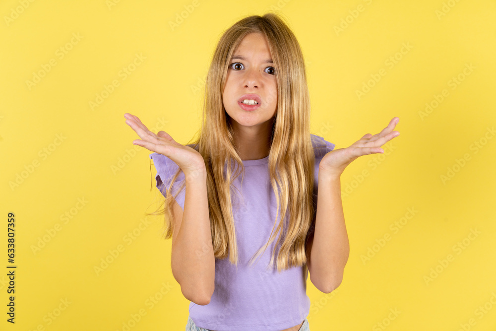 Indignant blonde kid girl wearing violet T-shirt over yellow studio background gestures in bewilderment, frowns face with dissatisfaction. - obrazy, fototapety, plakaty 