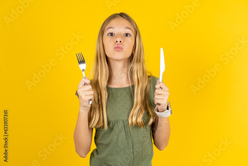 hungry blonde kid girl wearing green T-shirt over yellow studio background holding in hand fork knife want tasty yummy pizza pie