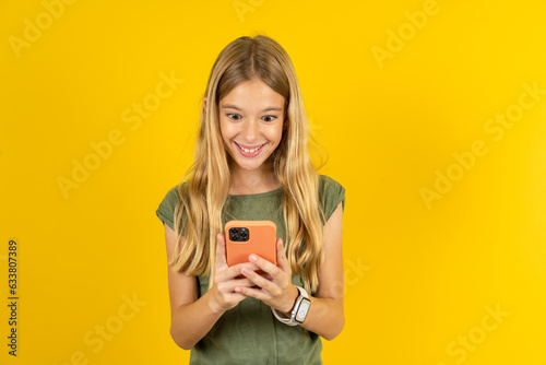 blonde kid girl wearing green T-shirt over yellow studio background using mobile phone chatting free time .
