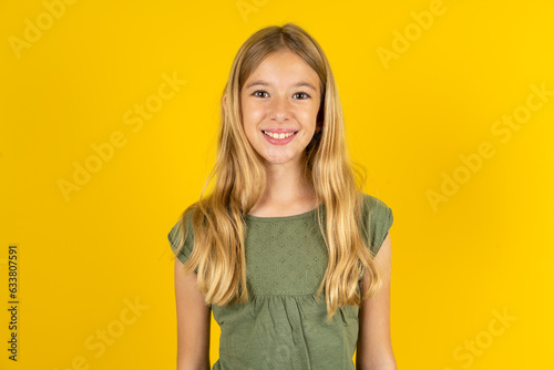 blonde kid girl wearing green T-shirt over yellow studio background with nice beaming smile pleased expression. Positive emotions concept