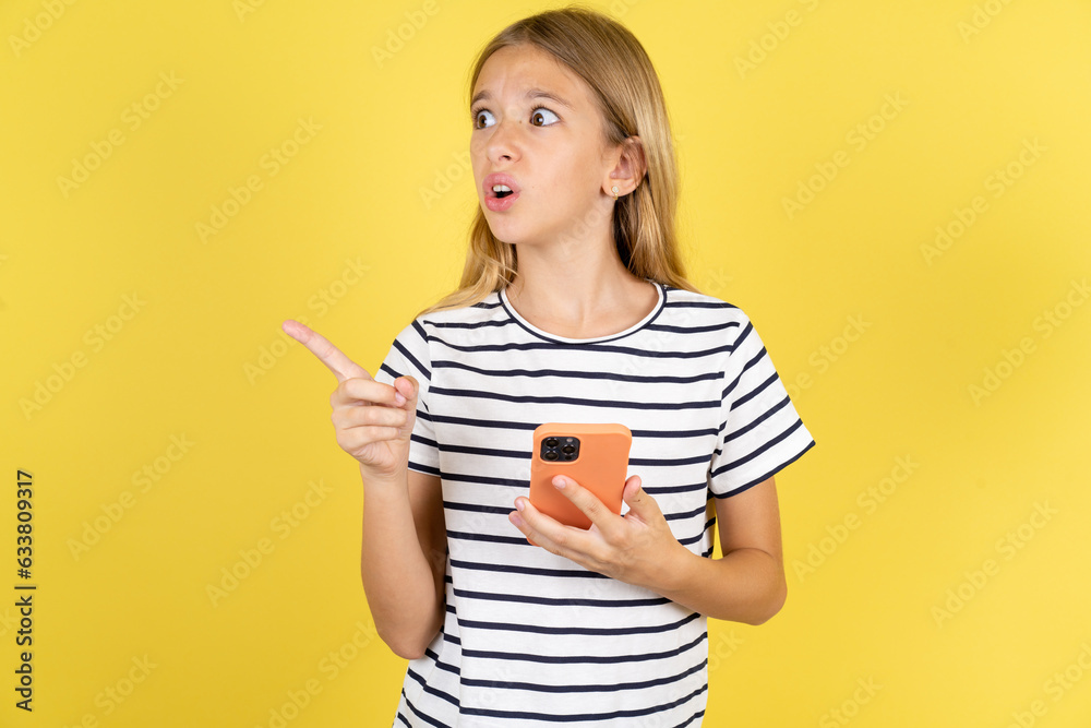Stunned young beautiful blonde kid girl over yellow studio background points sideways right copy space, recommends product, sees astonishing thing Stunned young beautiful blonde kid girl over yellow studio background points sideways right copy space, recommends product, sees astonishing thing