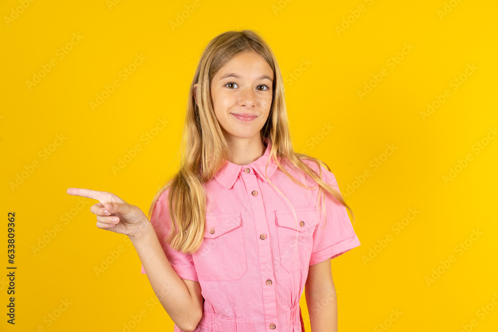 young beautiful blonde kid girl over yellow studio background points to side on blank space demonstrates advertisement. People and promotion concept young beautiful blonde kid girl over yellow studio background points to side on blank space demonstrates advertisement. People and promotion concept