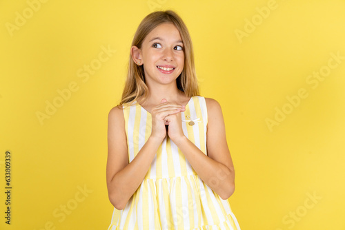 Happy young beautiful blonde kid girl over yellow studio background anticipates something awesome happen  looks happily aside  keeps hands together near face  has glad expression.