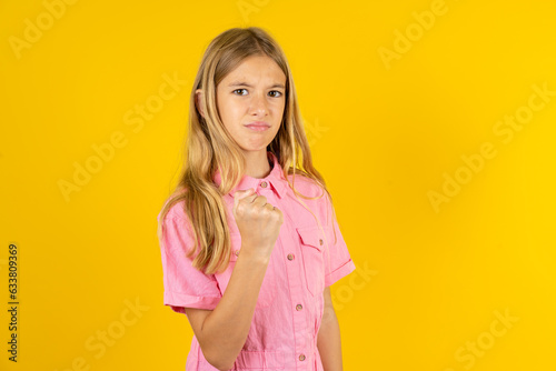 young beautiful blonde kid girl over yellow studio background shows fist has annoyed face expression going to revenge or threaten someone makes serious look. I will show you who is boss