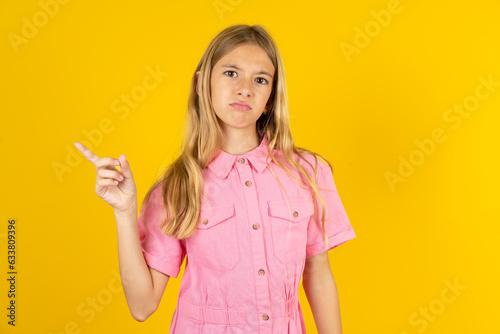 Serious young beautiful blonde kid girl over yellow studio background smirks face points away on copy space shows something unpleasant. Look at this advertisement. Big price concept