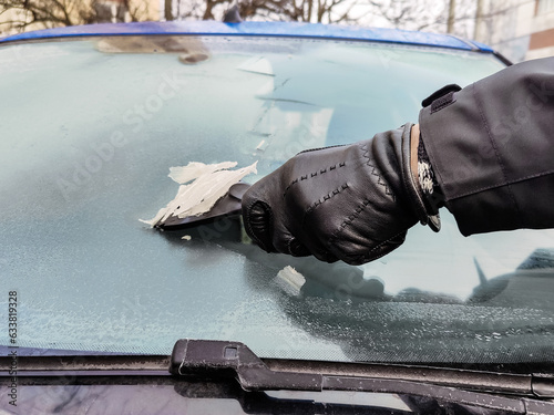 Cleaning car windows in winter. Man scrapes hoarfrost with a plastic scraper from the windshield of a blue car. Hand in a black leather glove in the cold. Close-up. Selective focus.