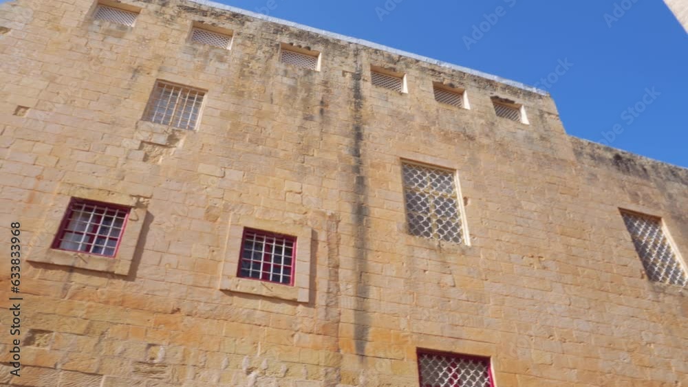 Mdina, Malta POV day low angle pan to traditional limestone low-rise buildings with windows and balconies inside the medieval Silent City citadel, under a blue sky. Mdina, Malta POV day low angle pan to traditional limestone low-rise buildings with windows and balconies inside the medieval Silent City citadel, under a blue sky.