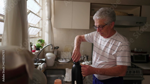 Mature man serving coffee with thermos bottle into glass by kitchen sink. Authentic domestic scene of retired older person starting the day ritual morning photo