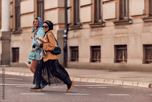 Couple woman one wearing a hijab and a modern yet traditional dress, and the other in a blue dress and scarf, walking together through the city at sunset. One carries a bouquet and bread, while the