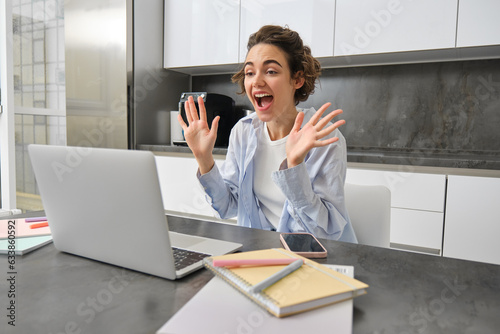 Friendly smiling woman sists at home, waves hands at laptop camera, says hello, joing online meeting, connects to video chat photo