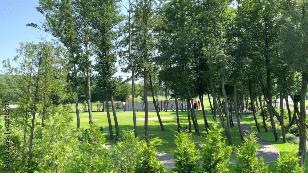 View through tall leafy trees swaying in the wind on a sunny day to a modern golf club and a green golf course where people are playing. Active recreation is a summer sport. View through tall leafy trees swaying in the wind on a sunny day to a modern golf club and a green golf course where people are playing. Active recreation is a summer sport.