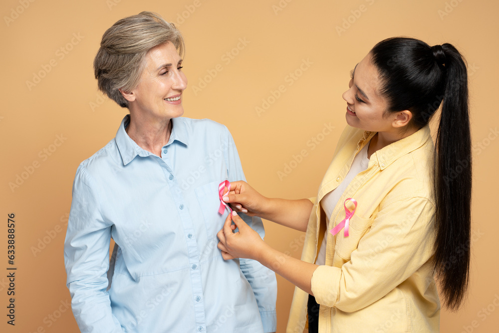 Portrait of smiling confident women attach pink ribbon looking each other isolated on beige background. Health care, support, solidarity, prevention. Breast cancer awareness month concept Portrait of smiling confident women attach pink ribbon looking each other isolated on beige background. Health care, support, solidarity, prevention. Breast cancer awareness month concept