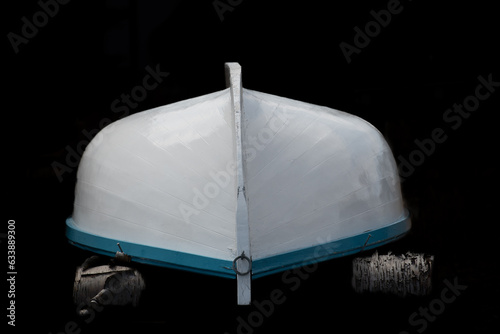 A white wooden traditional Rodney boat or small fishing vessel with blue trim and a rib down the middle on wood blocks. The rowing boat has a shiny white coat of paint and the background is black. 