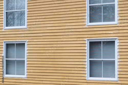 The exterior wall of a vintage yellow wooden house with four double hung window. The wall is pine cape cod siding with horizontal boards. The glass windows have white trim and white blinds hanging. 