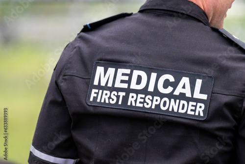 A close-up of a Caucasian male emergency health medical first responder or paramedic wearing a black uniform with grey letters. The ambulance attendant is wearing a short sleeve shirt with lettering.