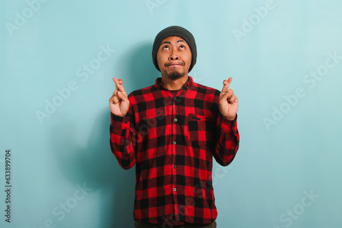 A hopeful young Asian man with a beanie hat and a red plaid flannel shirt makes a finger crossed gesture, looking up at empty copy space, and makes a wish while standing against a blue background