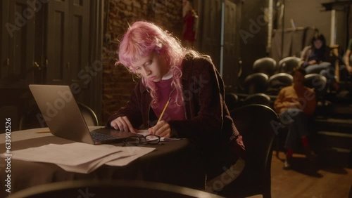 Young female playwright with pink hair using laptop, writing notes and posing for camera with smile on theater stage. Medium long shot, video portrait