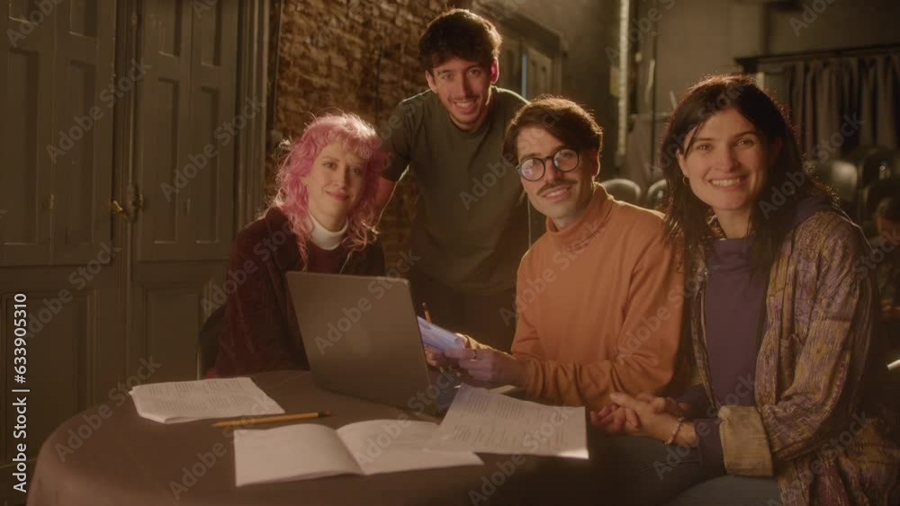 Group portrait of drama actors and director posing together for the camera and smiling at table with laptop papers on theater stage Group portrait of drama actors and director posing together for the camera and smiling at table with laptop papers on theater stage