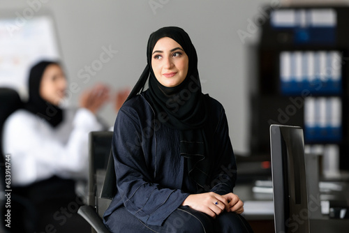 Sitting Arab Emirati woman at office with Arabic colleagues, around looking far away. Arabic employee wearing Hijab Abaya.