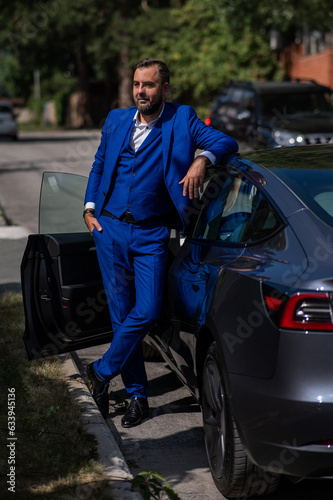 Caucasian bearded man in a blue suit gets out of a black electro car in the countryside in summer. © Михаил Решетников