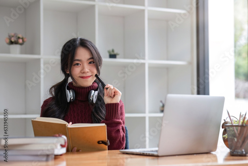 Asian female student studying online using headphones and laptop taking notes in notebook sitting at home table relaxing and changing places to school relax.