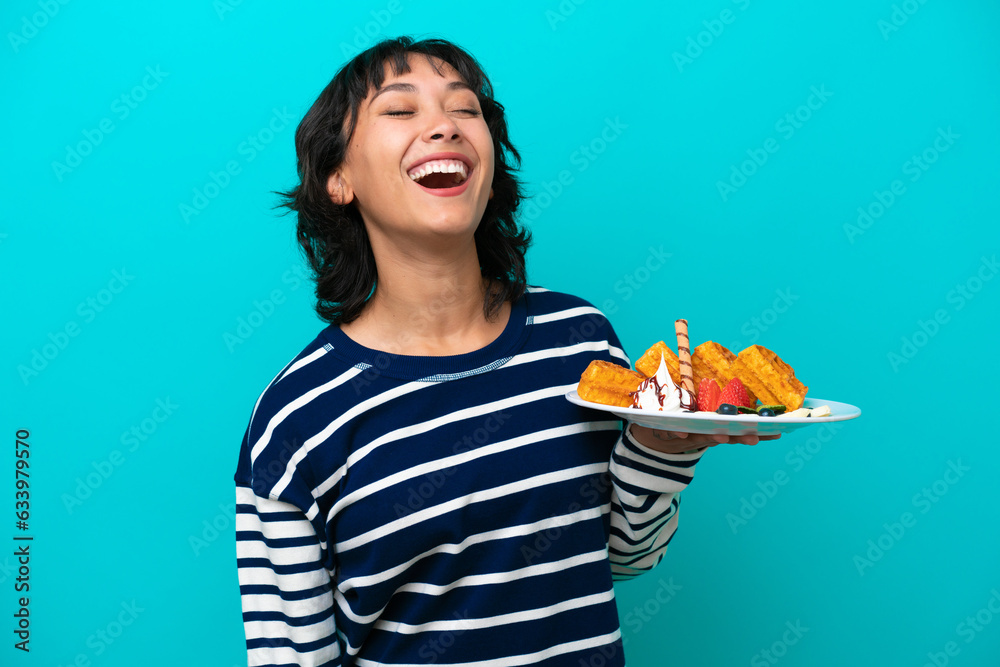 Young Argentinian woman holding waffles isolated on blue background laughing Young Argentinian woman holding waffles isolated on blue background laughing