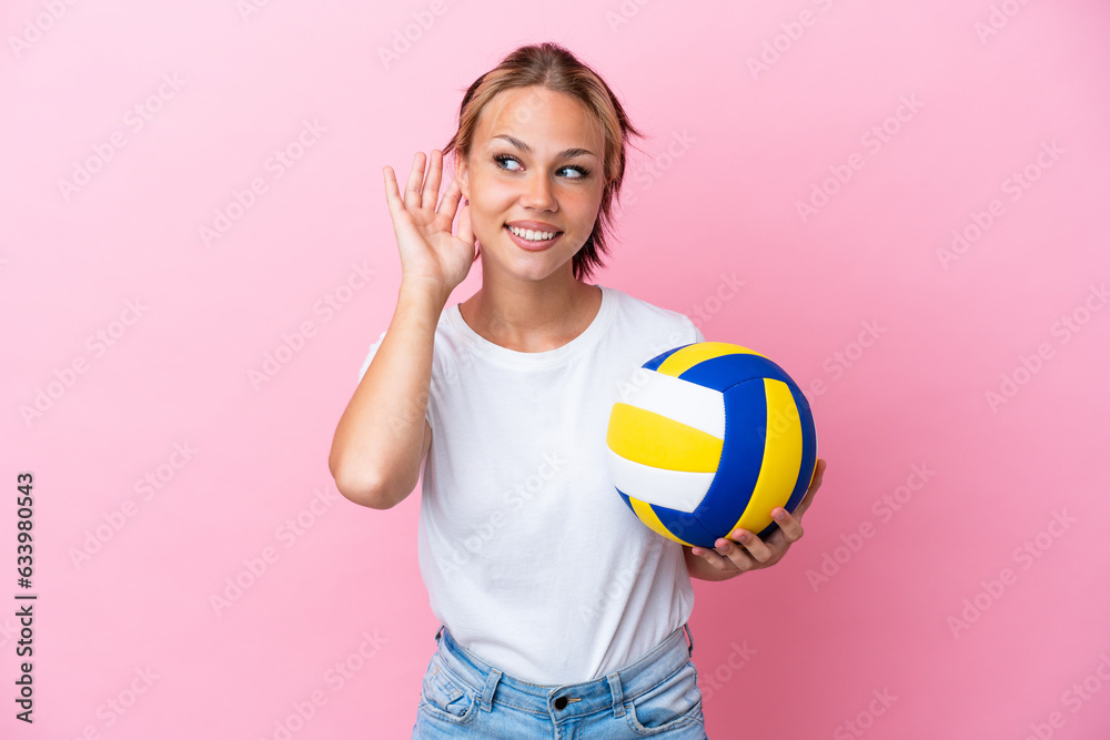 Young Russian woman playing volleyball isolated on pink background listening to something by putting hand on the ear