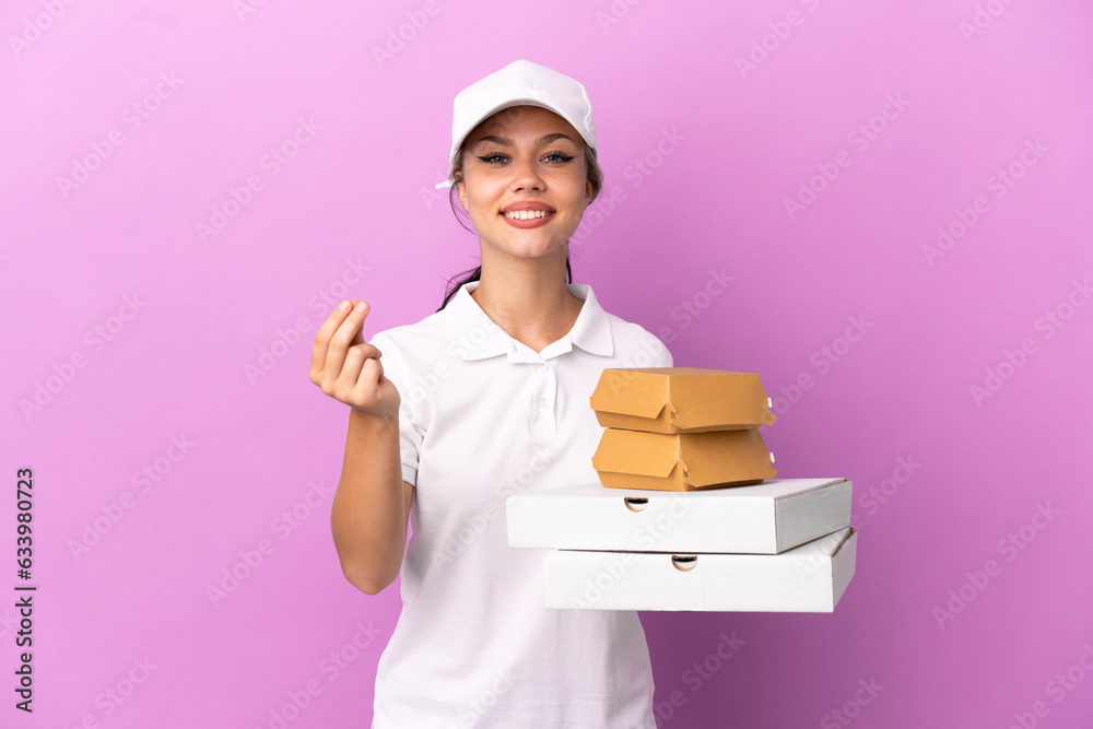 Pizza delivery Russian girl with work uniform picking up pizza boxes and burgers isolated on purple background making money gesture Pizza delivery Russian girl with work uniform picking up pizza boxes and burgers isolated on purple background making money gesture