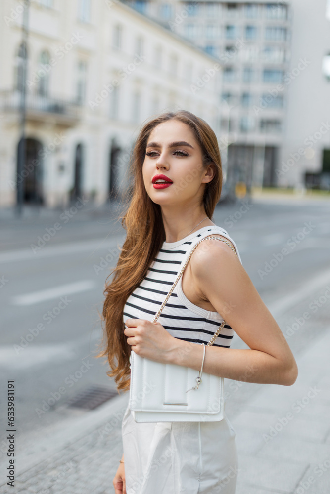 Urban beautiful young fashionable woman model in a trendy striped top and skirt with a white stylish bag walks in the city