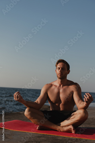 Confident young shirtless man practicing yoga while meditating on exercise mat near the sea © gstockstudio