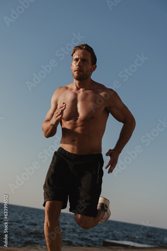 Confident young shirtless man jogging outdoors with the sea on background