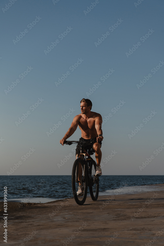 Fototapeta premium Handsome young shirtless man enjoying summer morning while cycling seaside