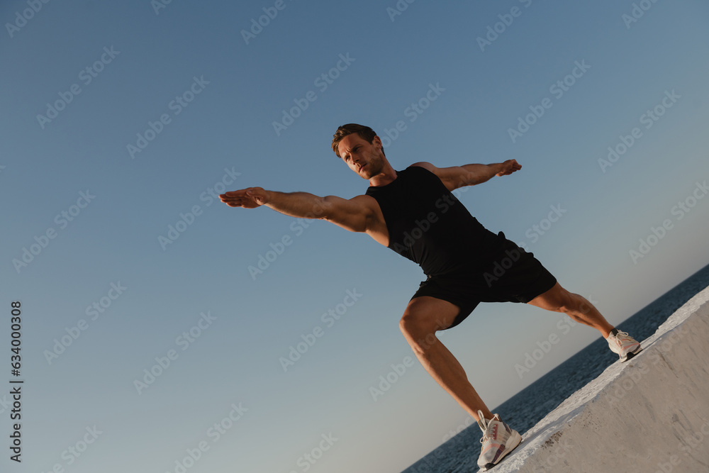 Low angle view of young male athlete doing stretching exercises with the sea view on background