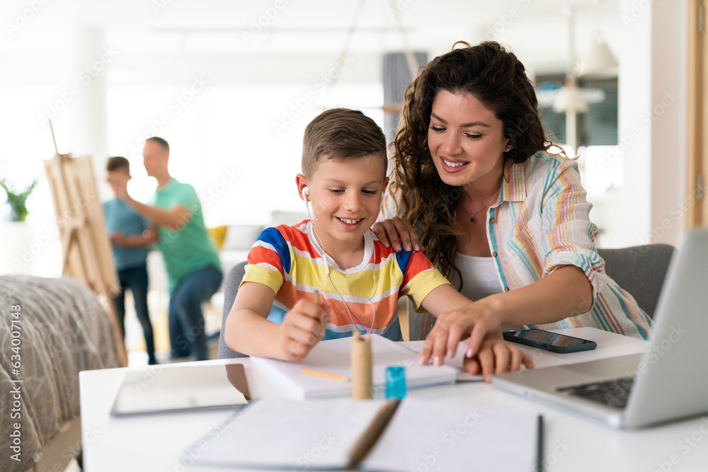 Patient and caring mother helping her son to finish his school work using laptop and tablet. They are writing it in the notebook while father and younger boy paint on canvas on an easel in background.