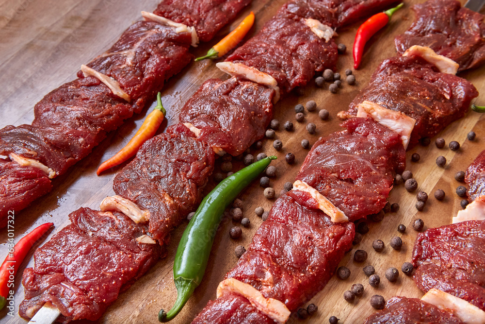 Metal skewers with raw uncooked beef meat for frying on the wooden board with chili peppers and pepper near it, close-up perspective view shallow depth of field. Meat, peppercorns and pepper in focus Metal skewers with raw uncooked beef meat for frying on the wooden board with chili peppers and pepper near it, close-up perspective view shallow depth of field. Meat, peppercorns and pepper in focus