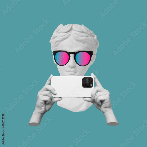 Antique statue's head in sunglasses holding mobile phone with photo camera in hands taking picture on color background. 3d trendy creative collage in magazine style. Contemporary art. Modern design photo