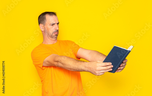 Hispanic man with a beard in his 40s pushing away the book he is trying to read since he cannot read up close, loss of eye vision due to age. Isolated on yellow studio background.