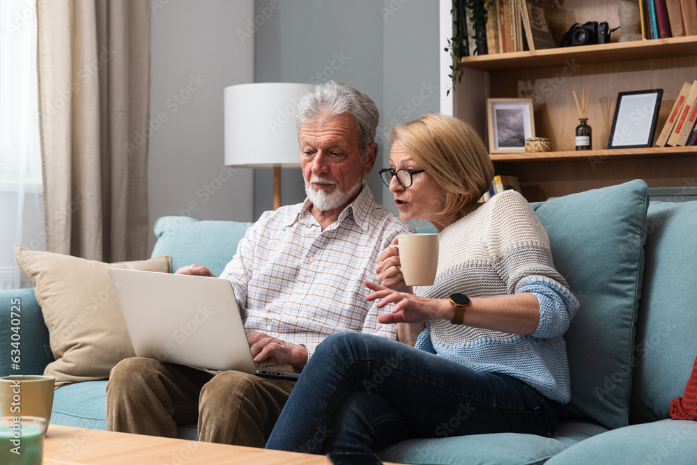 Elderly retired couple sitting at home surfing the internet on a laptop looking to buy a camper and sell their home to travel the world. Senior man and woman using computer to find travel destinations