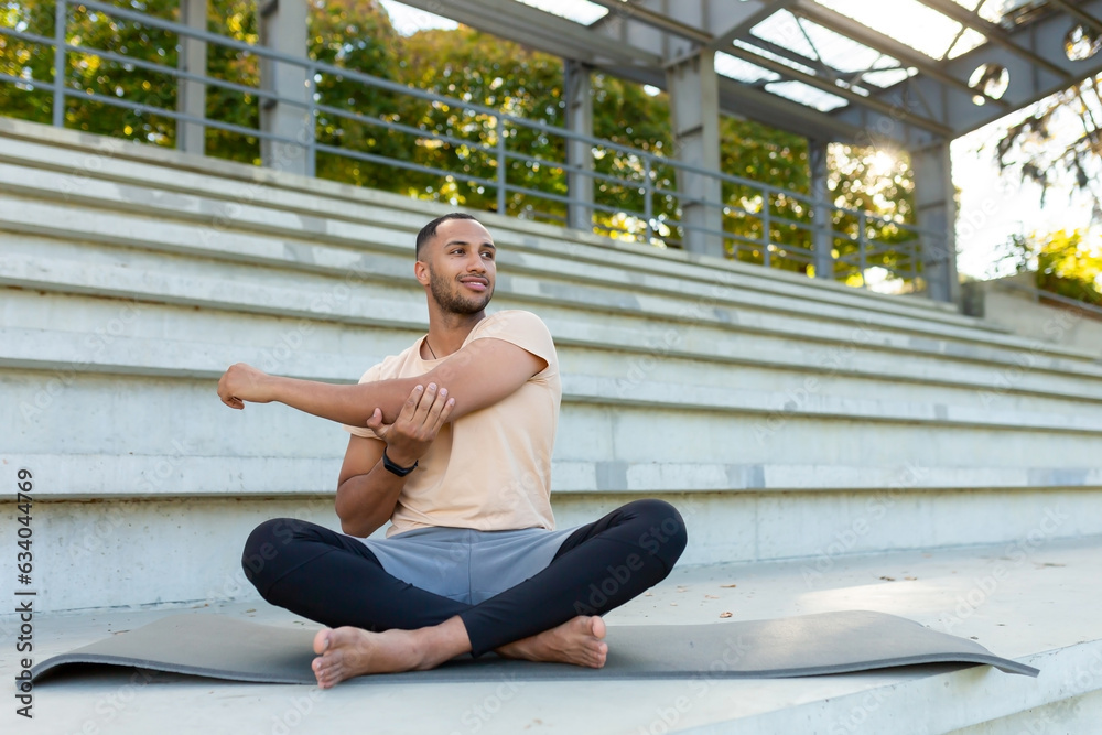 A young African American man is playing sports at the stadium. Sits on a mat, performs an exercise, stretches, does yoga