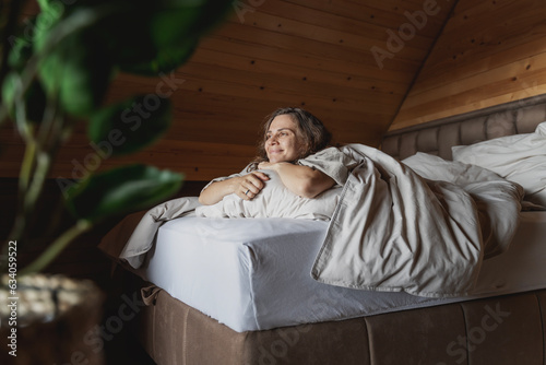 Young happy caucasian woman lying on bed with beige pastel linens hugging pillow and enjoying comfortable rest in log house © olezzo
