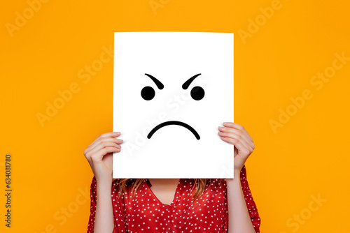 angry emoji. Girl holds a yellow paper poster with emoticon isolated over orange background