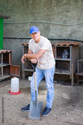 A male farmer leans on a shovel in an open rabbit farm.