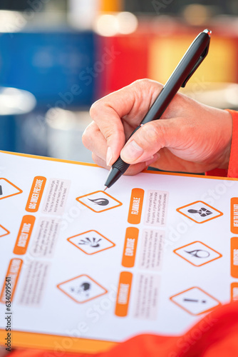 Action of safety officer is using a pen to checking on the hazadous material symbol label form with the chemical barrel as blurred background. Safety industrial working scene concept. Selective focus. photo